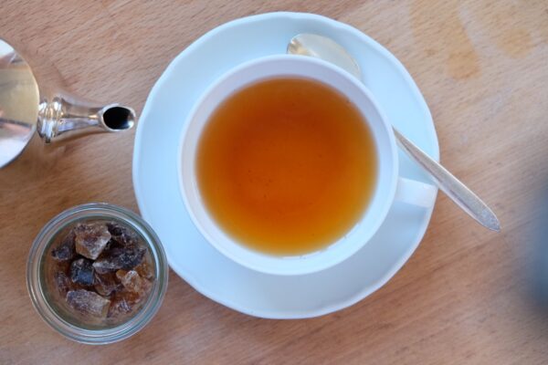 98F6D899-0152-45AD-87BE-DE5FE28501BD-2296-0000017C256CF2E5-2.jpg Eine Tasse SYLTER RUMTOPF TEEMISCHUNG mit Untertasse und Löffel steht neben einem Glas Kandiszucker und einer Teekanne auf der Holzfläche.