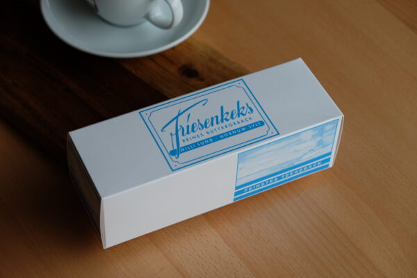 DSCF0114-scaled-1.jpg Eine Schachtel FRIESENKEKSE 240 g Butterkekse steht auf einem Holztisch neben einer weißen Tasse auf einer Untertasse.