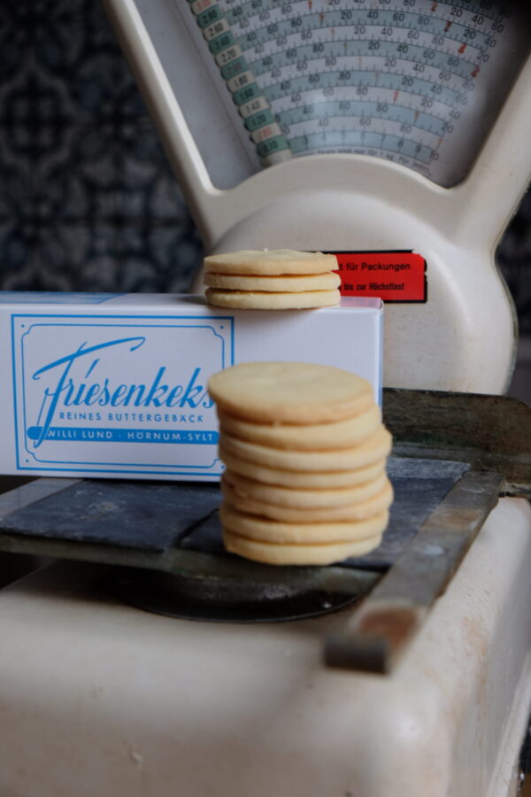 DSCF0118-scaled-1.jpg Ein Stapel runder Butterkekse mit der Aufschrift „FRIESENKEKSE 240g“ steht elegant auf einer Vintage-Waage, im Hintergrund ist eine hübsche Keksdose zu sehen.