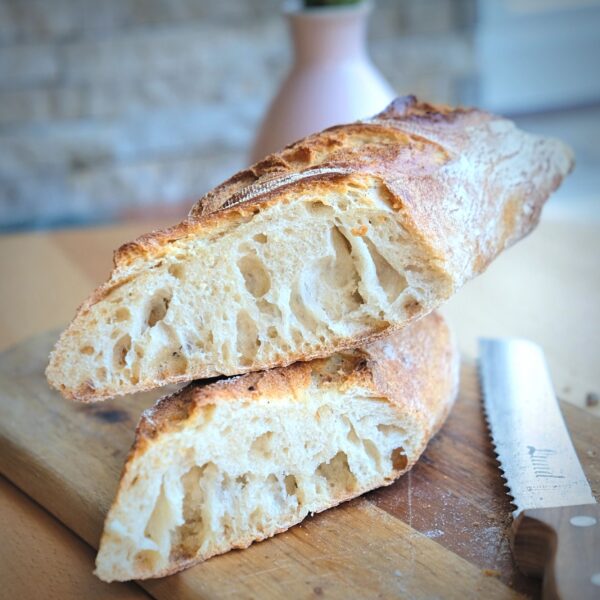 Dinkel_baguette_Lund_Sylt-2-3.jpg Zwei rustikale Hälften BIO DINKEL-BAGUETTE-Brotes aus kontrolliert biologischem Anbau liegen auf einem Holzbrett, daneben liegt ein gezacktes Messer.