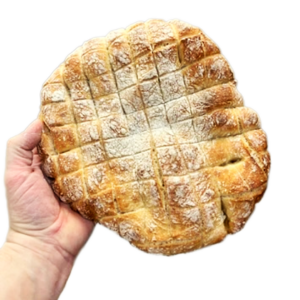 Eine Hand hält einen runden Laib BIO DINKEL FONDUE BROT, der eine knusprige Oberfläche mit einem Kreuzmuster aufweist.