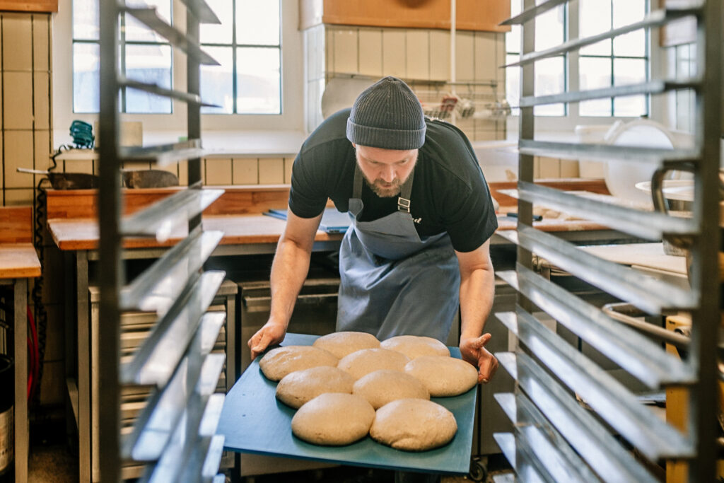 Ein Bäcker mit Schürze und Mütze legt ein Tablett mit Teigkugeln auf ein Gestell in einer Bäckereiküche, in der die Kunden Brot vorbestellen können, um frisches, leckeres Brot zu bekommen.