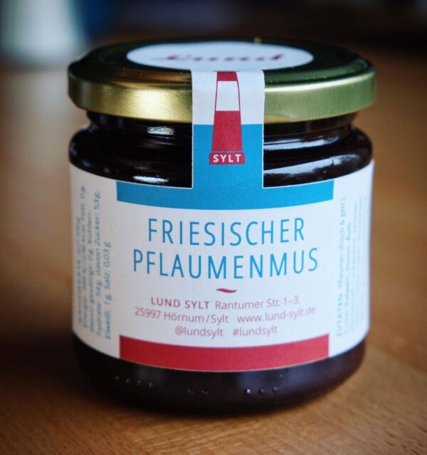 Sylter-pflaumenmus171222.jpg Ein Glas „LUNDS SYLTER PFLAUMENMUS“ mit einem rot-weißen Etikett und einem goldenen Deckel vor einem Holztischhintergrund.