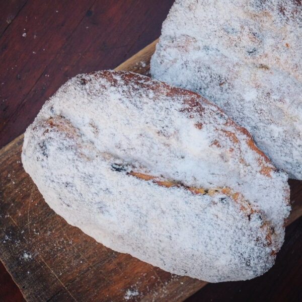 Sylter-stollen-Sylter-Milch-lund-sylt-online-shop-1.jpg Zwei Laibe SYLTER BUTTERSTOLLEN, mit Puderzucker bestäubt, liegen auf einer Holzoberfläche und laden Sie ein, ihre reichhaltige, buttrige Köstlichkeit zu genießen.