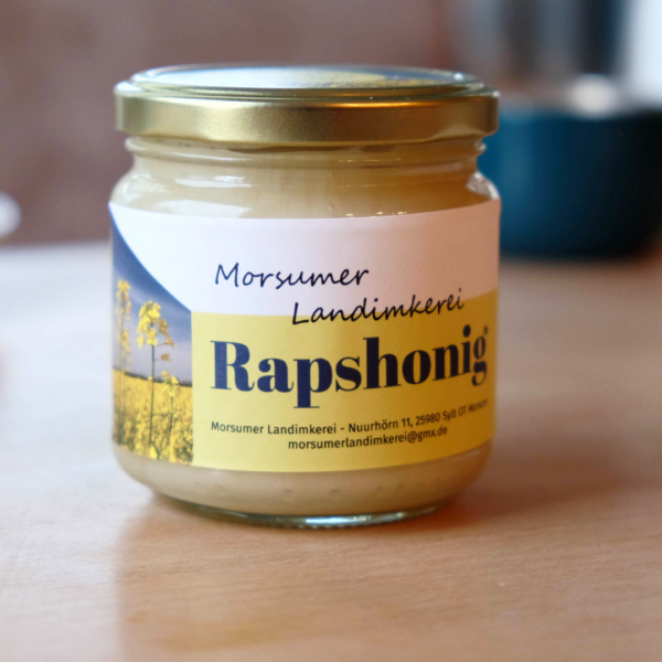 SylterRapshonig.png Auf einem Holztisch steht ein Glas Sylter Rapshonig, auf dem Etikett ist ein leuchtendes Feld gelber Blumen abgebildet.