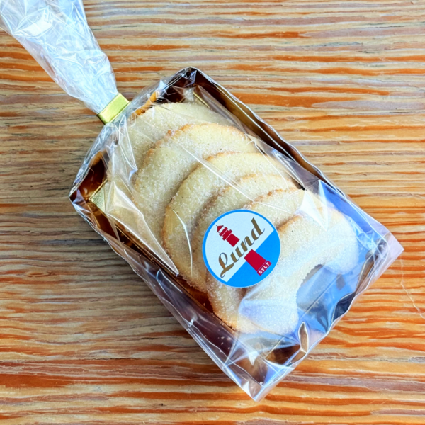 Tahiti-Vanille-Kipferl-Kipfel-Lund-Sylt.png Die TAHITI-VANILLE KIPFEL Zuckerkekse sind in einer Klarsichtverpackung mit einem „Lund“-Leuchtturmetikett verpackt, das elegant auf einer Holzoberfläche präsentiert wird.