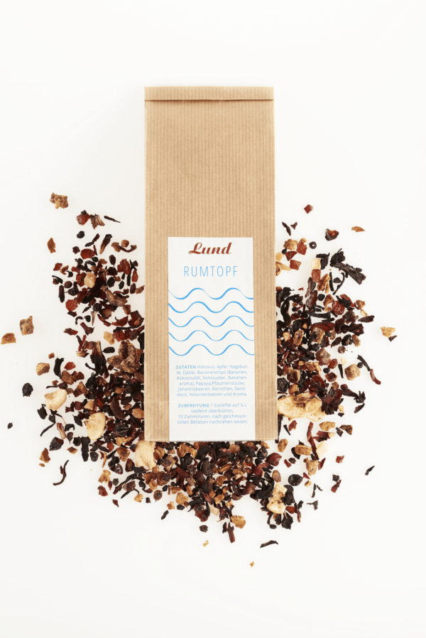 Tee-Rumtopf.png Eine 100 g Packung „SYLTER RUMTOPF TEEMISCHUNG“ ruht anmutig zwischen losen Teeblättern und bunten Trockenfrüchten und bietet einen köstlichen Aufguss.