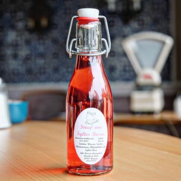 sylter-rosen-sirup-lund-sylt-1.jpg Eine Glasflasche SYLTER ROSENSIRUP mit Keramik-Bügeldeckel steht anmutig auf einem Holztisch im Innenbereich.