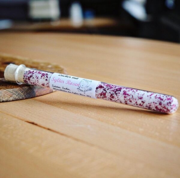 sylter-rosen-zucker-3.jpg Auf einem Holztisch steht ein Reagenzglas mit groben roten und weißen Kristallen, die an Sylter Aromen erinnern. Auf dem Etikett steht „SYLTER ROSENZUCKER“, was darauf hinweist, dass diese einzigartige Mischung von der Küste inspiriert ist.
